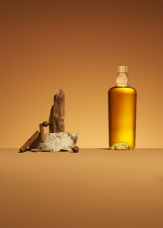 Botella de licor ámbar, modelo Malty, junto a una composición de cortezas, ramas de canela y avellanas sobre un fondo ocre.