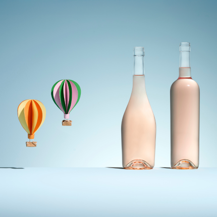 Deux bouteilles en verre blanc de formes différentes pour vin rosé, à côté de deux montgolfières colorées en papier sur un fond bleu pastel.