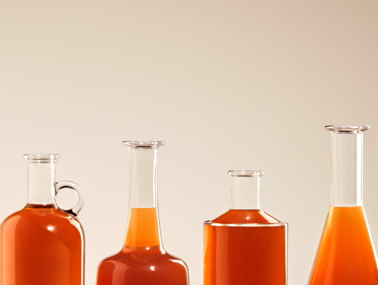 Gros plan sur quatre goulots de bouteilles en verre alignées, remplies d'un liquide orangé, présentant un assortiment de différentes bagues.